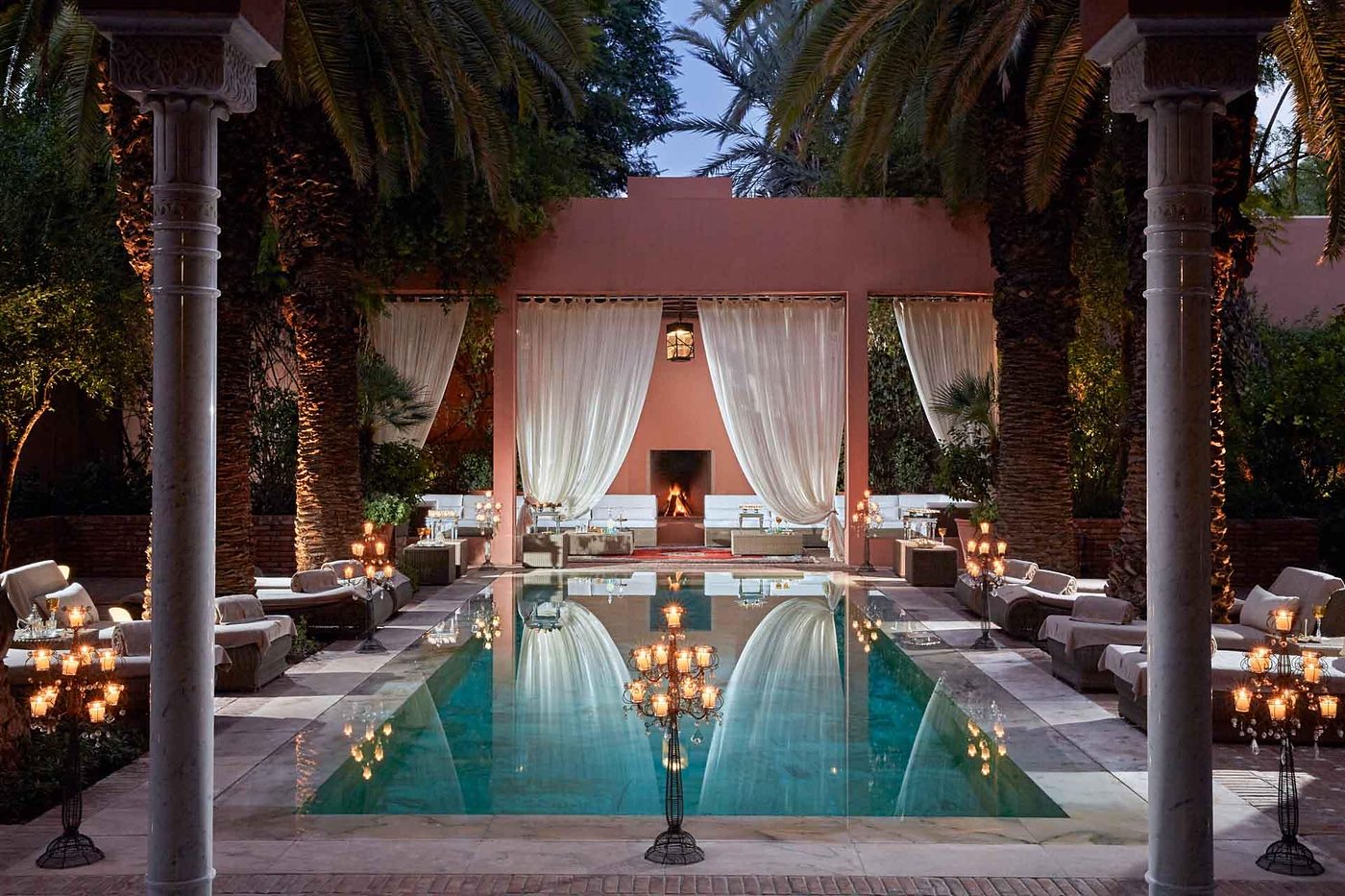 A candlelit pool courtyard at Royal Mansour Marrakech at dusk, framed by carved columns and palms