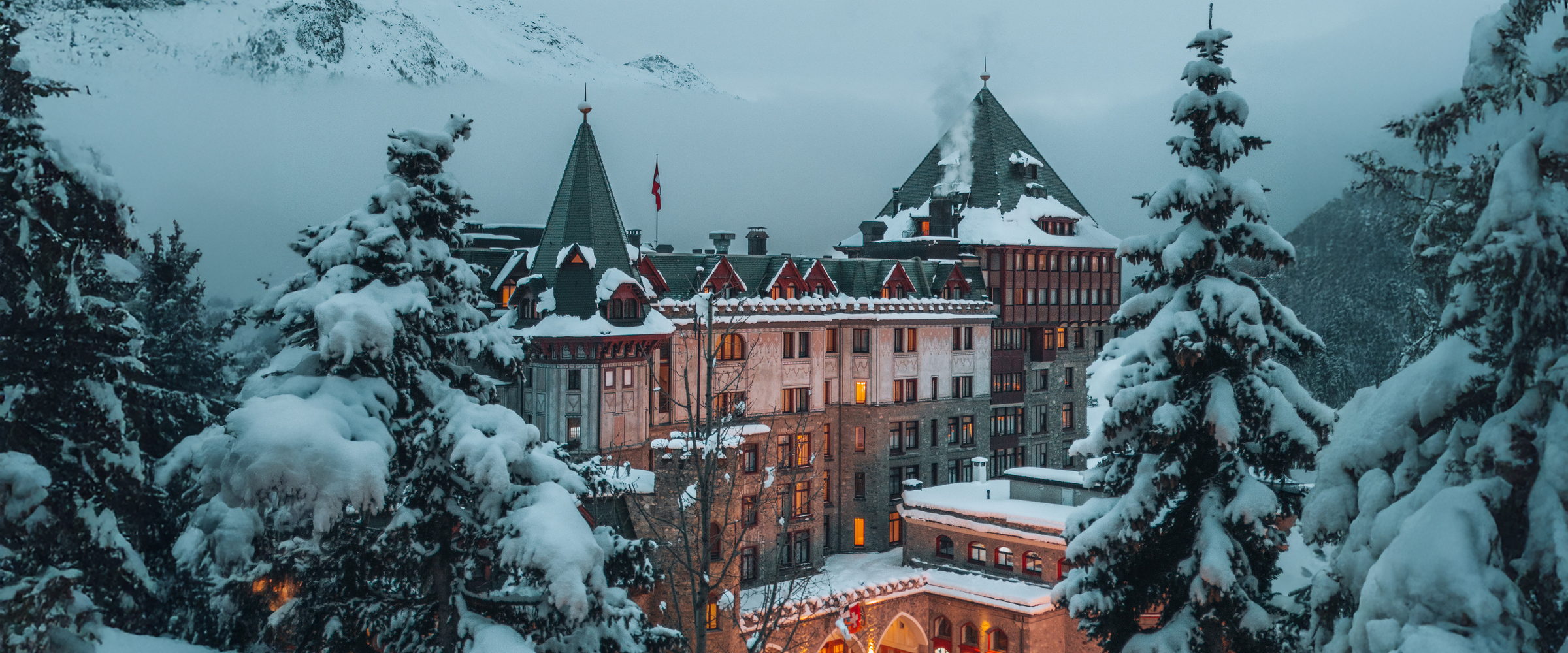 An alpine grand hotel lit at dusk in heavy snow, seen through snow-laden pines