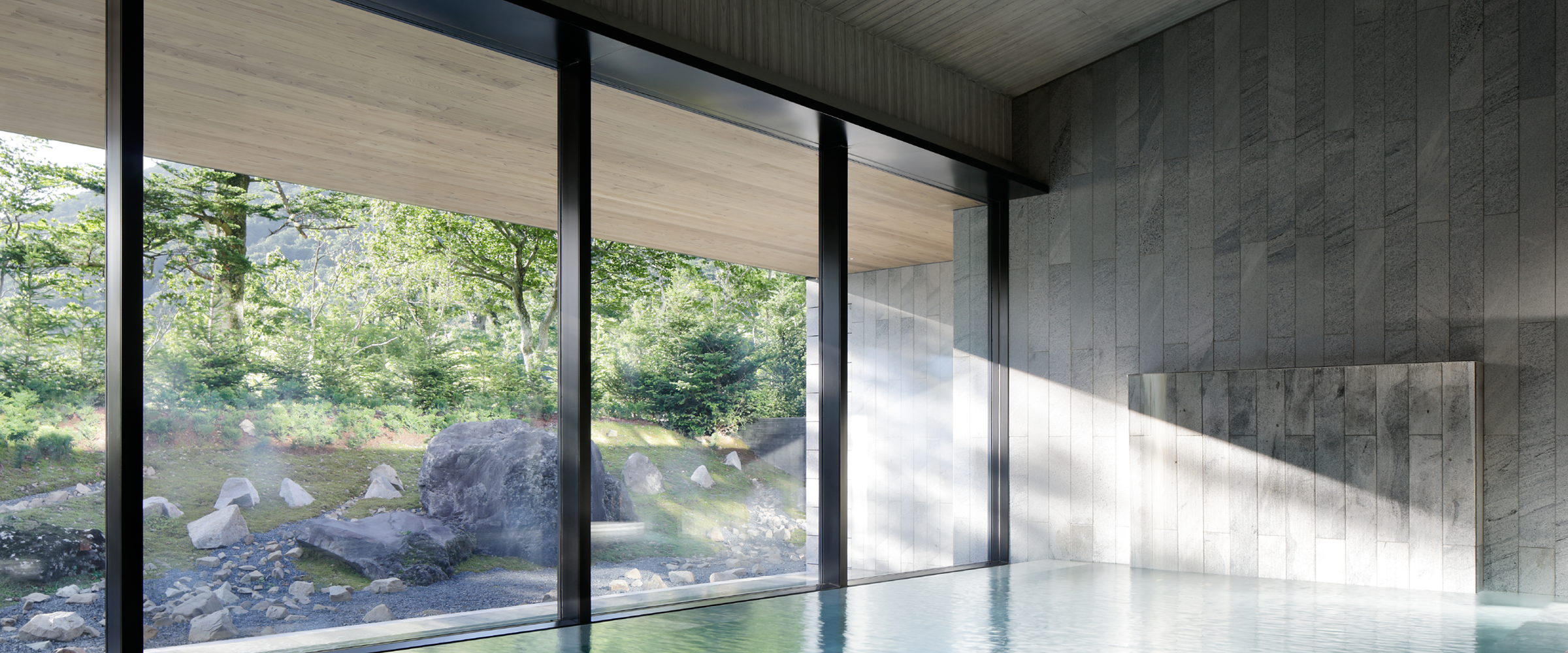 A stone-lined indoor onsen bath with full-height glazing onto a mossy rock garden and forest