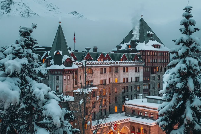 An alpine grand hotel lit at dusk in heavy snow, seen through snow-laden pines