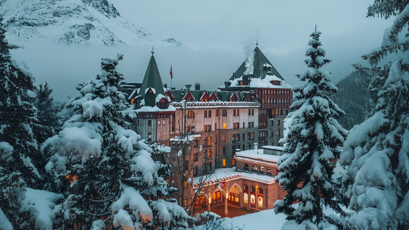 Badrutt's Palace Hotel lit at dusk in deep snow above St Moritz, framed by snow-laden pines